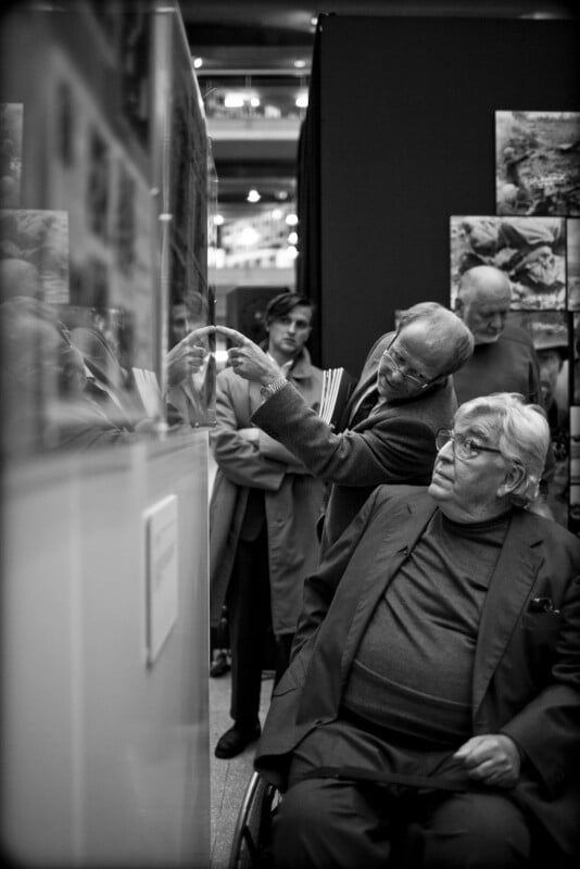 A man in a wheelchair looks on as another man points to a display behind glass. Three other people stand in the background, observing the exhibition. The scene is in black and white.