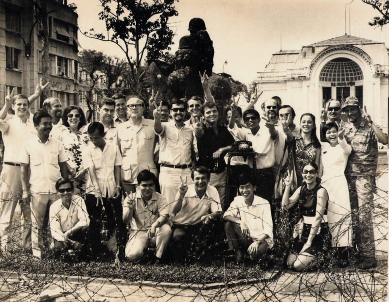A group of men and women, both Western and Asian, smile and gesture peace signs in front of a building and statue, with barbed wire in the foreground; the photo appears vintage, likely from the 1960s or 1970s.
