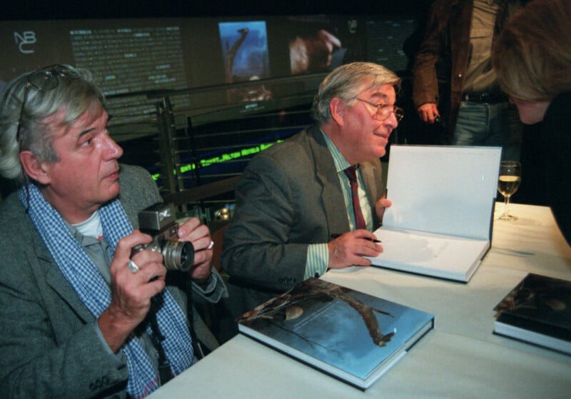 Two older men sit at a table with books; one holds a camera while the other, wearing glasses, signs a book for a woman. A wine glass and books with animal covers are on the table, and a busy backdrop is visible.