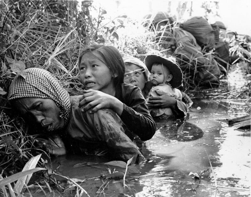 A woman and children wade through muddy water, holding onto each other, with soldiers in the background. Their expressions show fear and distress as they move through tall grass.