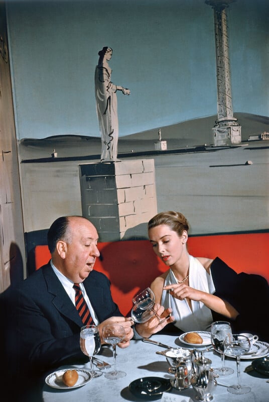 A man and a woman sit at a restaurant table with white tablecloths, glassware, and bread rolls. The man examines a wine glass while the woman gestures towards it. A mural of ancient statues and columns is on the wall behind them.