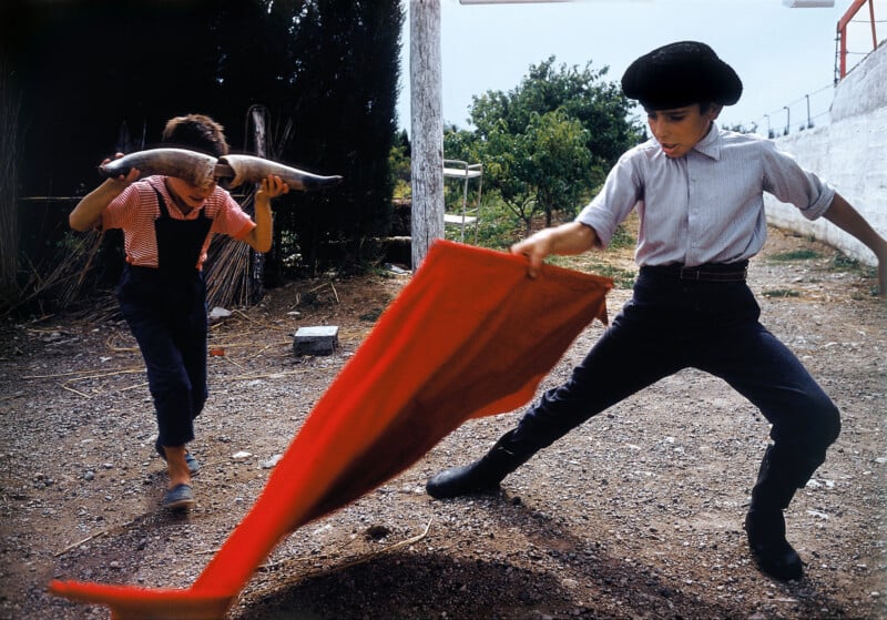Two children play matador and bull outdoors. One child waves a red cloth while dressed as a matador; the other wears horns and pretends to be a bull, charging at the cloth in a dirt-covered yard.
