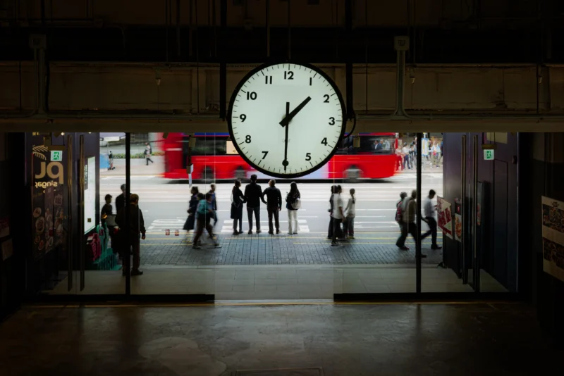 A large clock showing 10:10 hangs above a busy entrance, with people walking in and out. Outside, a red double-decker bus moves past the glass doors. The scene is urban and lively.