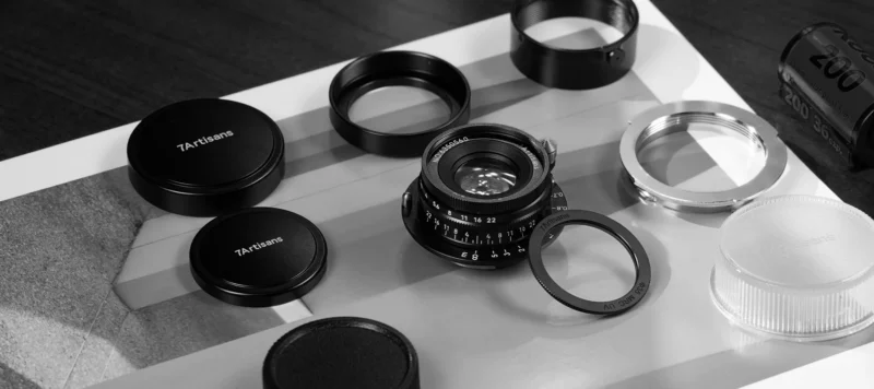 A black and white photo shows disassembled camera lens parts laid out on a table, including lens caps labeled "7Artisans," various rings, and other components arranged neatly.