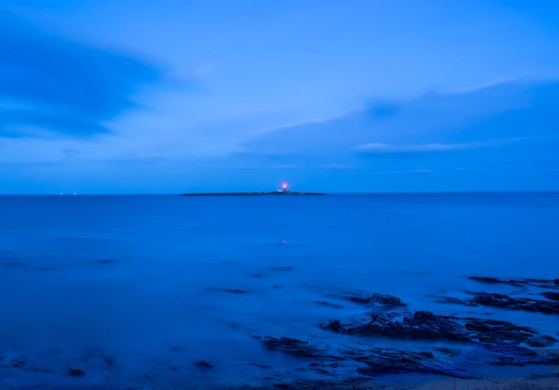 A calm seascape at dusk with a rocky shoreline in the foreground, smooth blue water, and a distant island featuring a lighthouse with a glowing red light under a blue sky.