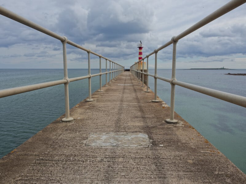 A long concrete pier with metal railings extends over the sea toward a distant red-and-white striped lighthouse under a cloudy sky.