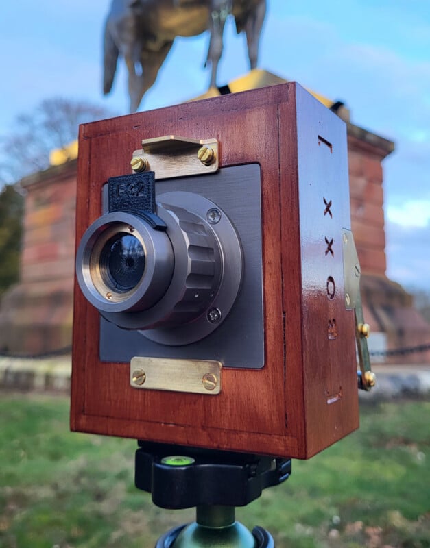 A wooden box camera with brass fittings and a metal lens is mounted on a tripod outdoors. A statue of a horse and rider on a stone pedestal is visible in the blurred background.