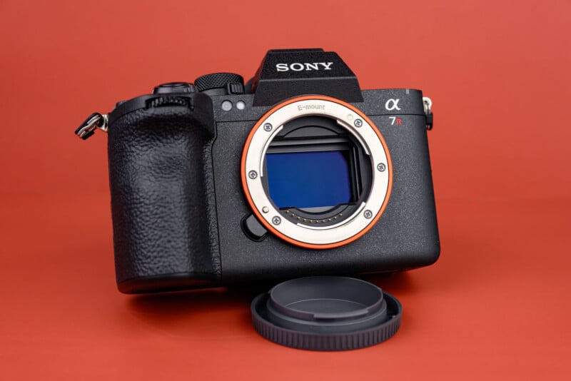 A Sony α7R mirrorless camera body with the lens removed, showing the sensor and lens mount, placed on an orange surface next to a detached lens cap.