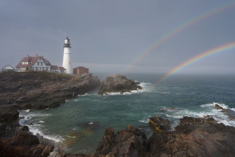 A lighthouse stands on a rocky coast beside a house, overlooking rough ocean waves. Two vivid rainbows arc across the misty sky above the water.