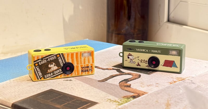 Two small toy cameras with Peanuts-themed designs, one yellow and one green, sit on a book near a window. The green camera features Snoopy and a tent; the yellow camera has a "Care-Free Vibe" label and Woodstock.