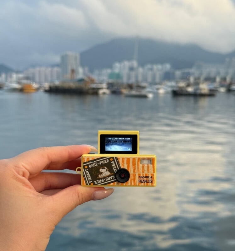 A hand holds a small yellow camera with a screen displaying a photo of a cityscape; in the background, there’s a harbor with boats and tall buildings under a cloudy sky and mountains.