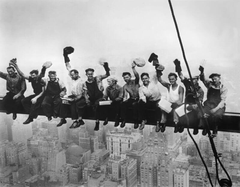 Eleven construction workers sit on a steel beam high above New York City, eating lunch and waving their hats, with skyscrapers visible far below in the background.