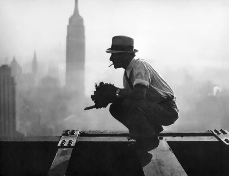 A man wearing a hat and smoking a cigarette crouches on a steel beam high above a city, with the Empire State Building visible in the misty background. He holds a camera, suggesting he is a photographer.
