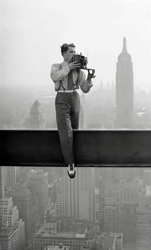 A man in dress pants, suspenders, and a button-up shirt sits on a steel beam high above a city skyline, holding a vintage camera with the Empire State Building visible in the background.