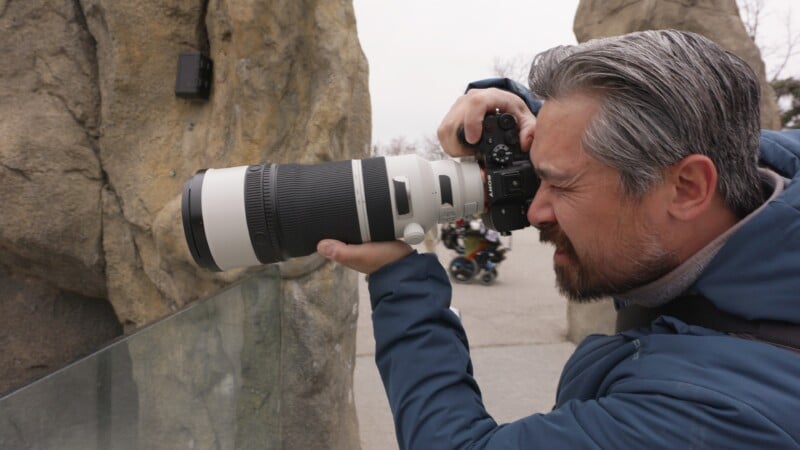 A man with gray hair and a beard uses a professional camera with a large telephoto lens, aiming it intently at something beyond a glass barrier, with rocky structures and blurred people in the background.