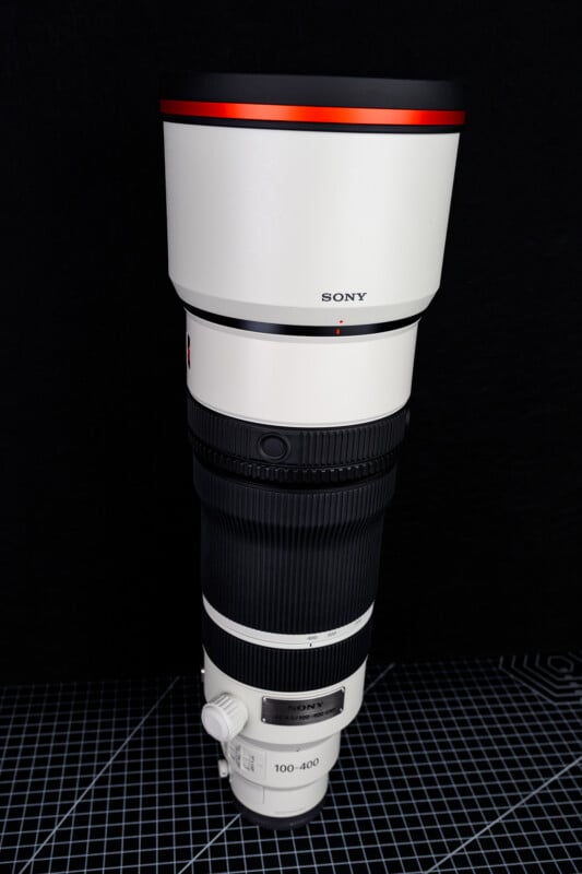 A large white Sony camera telephoto lens with a red ring near the top is standing upright on a grid-patterned surface, against a black background.