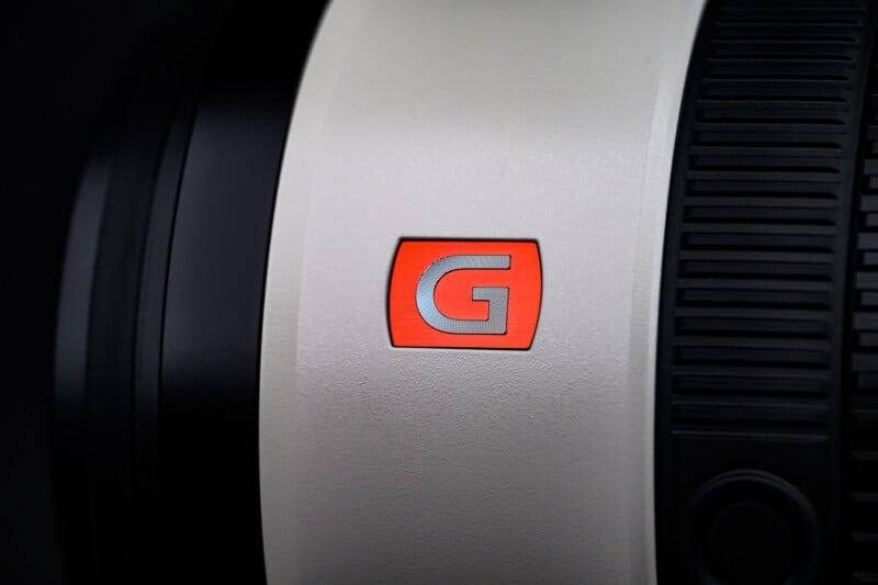 Close-up of a camera lens featuring a red "G" logo on a white background, with a textured black focus or zoom ring to the right.