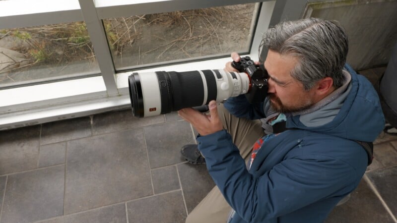 A man with gray hair and a beard, wearing a blue jacket, holds a professional camera with a large telephoto lens, aiming it upwards indoors near large windows.