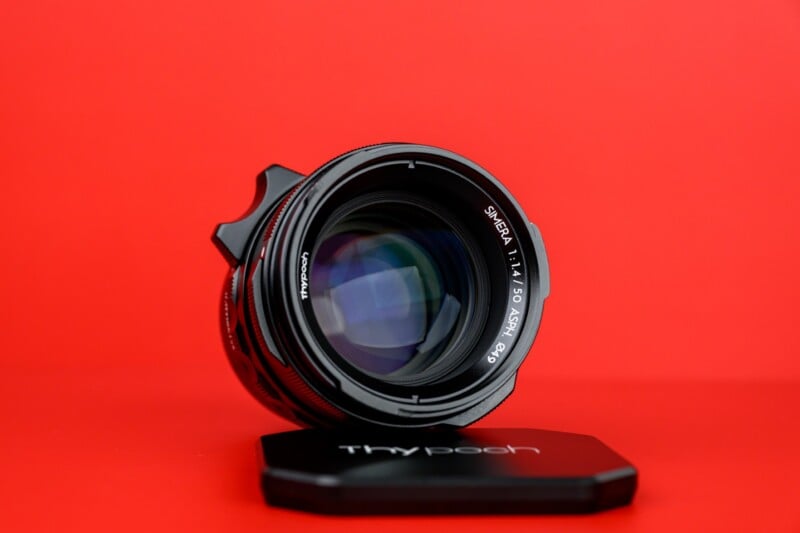 A camera lens with visible glass elements and white text markings is positioned upright on a red background, with its lens cap labeled "Thypoch" placed in front.