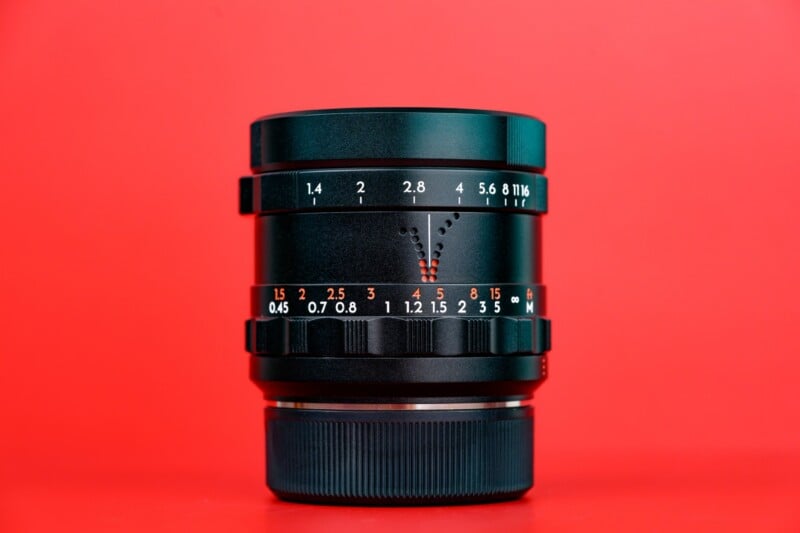 A black camera lens with aperture and focus distance markings in white and orange, displayed upright against a vibrant red background.
