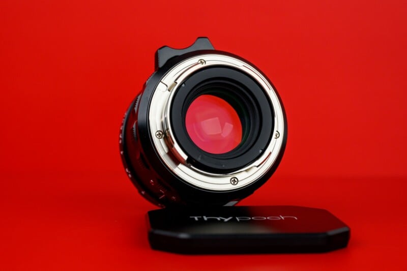 A close-up of a camera lens placed upright on a black lens cap with "Thypoch" written on it, set against a solid bright red background.