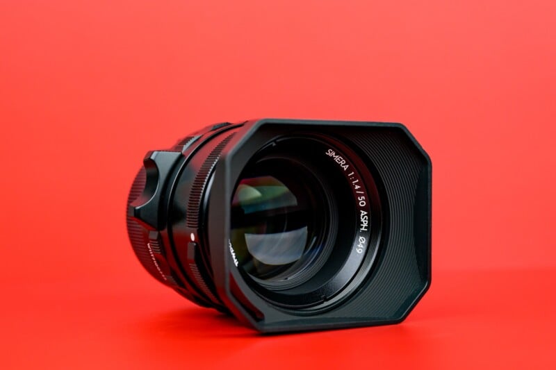A black camera lens with a lens hood is placed on a solid red surface, set against a red background. The lens markings read "SPHERAL 1:1/50 ASPH.