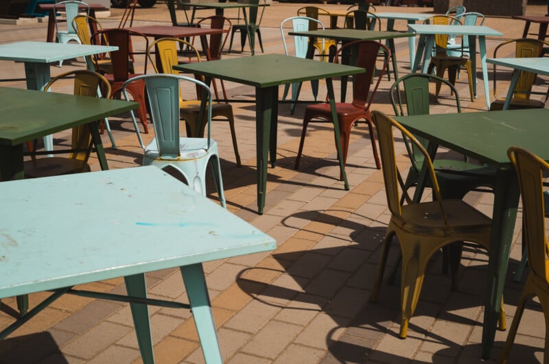 Outdoor seating area with empty, colorful metal chairs and rectangular tables on a paved surface, arranged in no particular order under bright sunlight.