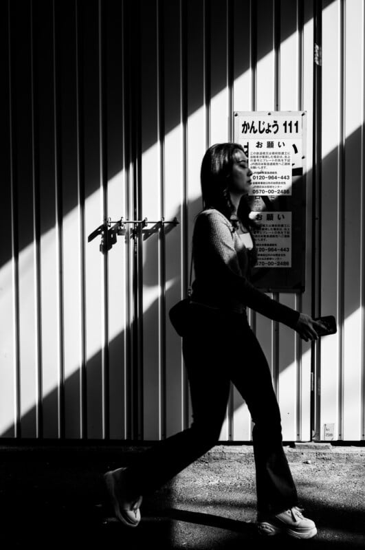 A woman walks briskly past a metal wall with dramatic diagonal shadows and a sign in Japanese. She is in profile, holding a phone, with light and shadow creating a striking contrast on the scene.