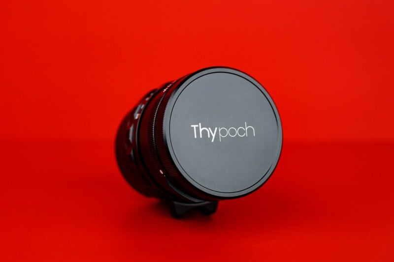 A camera lens with a cover labeled "Thypoch" is placed on its side against a bright red background.