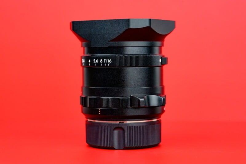 A black camera lens with a lens hood sits upright on a solid red background, showing aperture markings and manual adjustment rings.