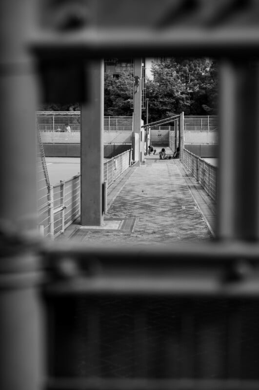 Black and white image showing a walkway framed by metal bars in the foreground. In the distance, two people sit on the ground near a fence, surrounded by urban structures and trees.