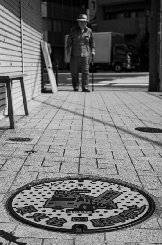A man with a cane walks on a city sidewalk in Osaka, Japan. The foreground shows a decorated manhole cover labeled "Osaka City." The photo is in black and white.