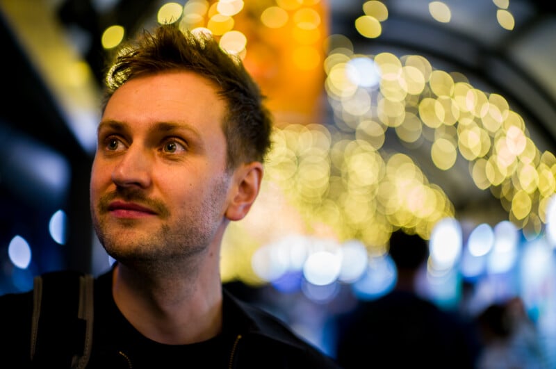 A man with short brown hair and stubble is looking slightly upward and to the side, standing under a canopy of warm, blurred string lights at night, creating a festive and dreamy atmosphere.