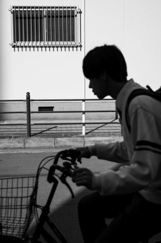 A silhouette of a person riding a bicycle in front of a white wall with a barred window and a metal railing, photographed in black and white.