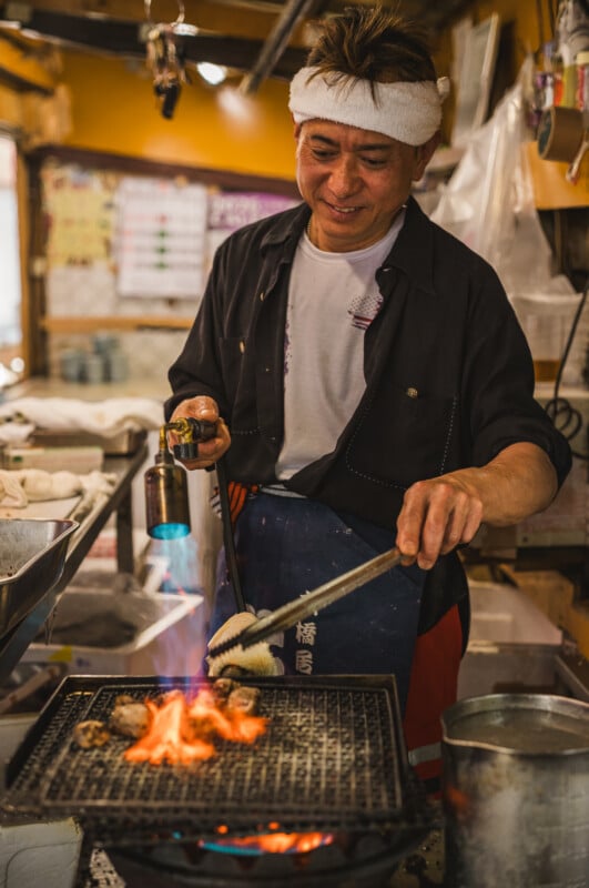 A smiling chef with a white headband uses tongs and a blowtorch to grill food over an open flame in a cozy, warmly lit kitchen. Various kitchen tools and ingredients surround him.