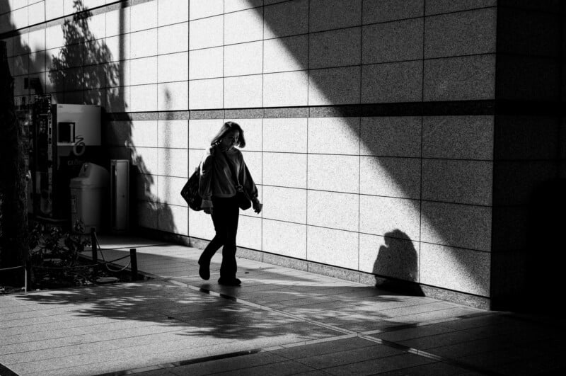 A person walks alone on a sunlit sidewalk next to a textured wall, casting a distinct shadow. The scene is in black and white, with sharp contrasts between light and dark areas.
