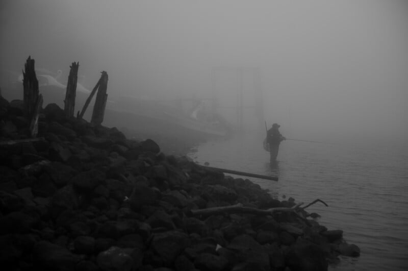 A person stands in shallow water fishing with a rod on a foggy shoreline, surrounded by rocks and driftwood, with boats barely visible in the background.