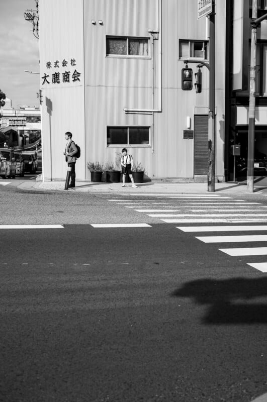 A black-and-white photo of a man and a child standing on a sidewalk at a crosswalk near a building with Japanese writing, waiting for the traffic signal to change. Shadows and sunlight create contrast on the street.