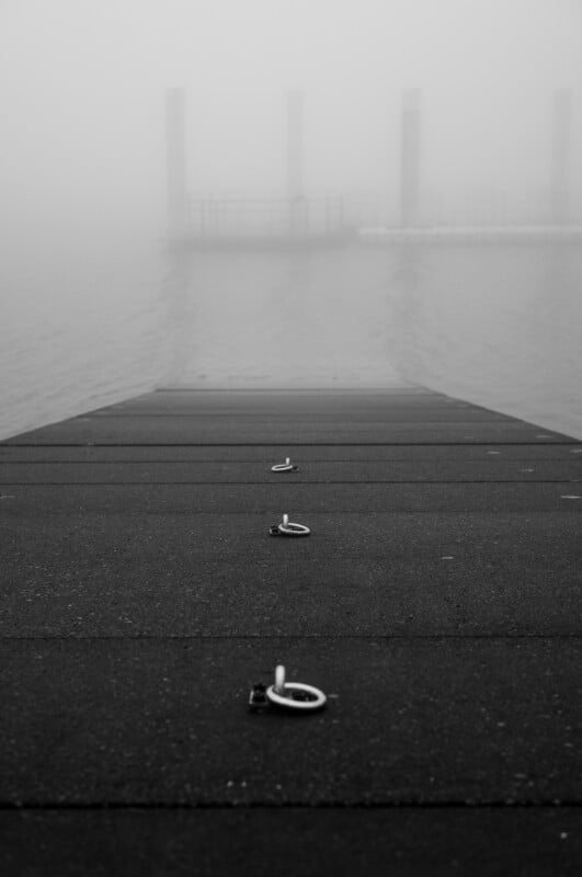 A foggy scene showing the end of a dock with metal hooks on its surface, leading out over water. In the background, faint outlines of pillars and a floating platform are barely visible in the mist.