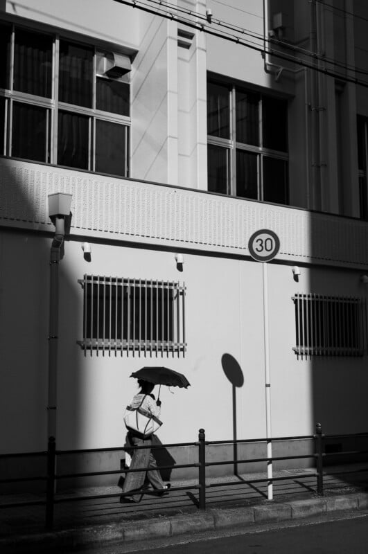 A person walks along a sunlit street holding an umbrella, casting a shadow on a white building with barred windows and a "30" speed limit sign. The scene is in black and white.