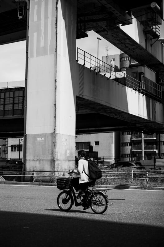 A person rides a bicycle along a city street under large elevated concrete structures, with tall buildings in the background. The image is in black and white.