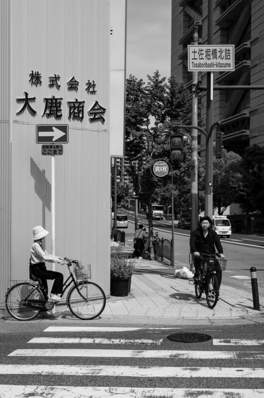 Two people ride bicycles across a crosswalk in an urban area. One cyclist wears light clothing and a hat; the other wears dark clothing. Japanese signs and buildings are visible in the background. The image is in black and white.