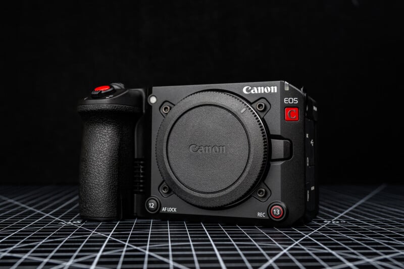 A Canon EOS camera body with a lens cap attached, featuring a red record button and grip, is displayed on a grid-patterned surface against a black background.