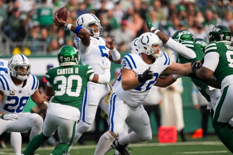 Een actiefoto van een American football- wedstrijd: een quarterback in een wit-blauwe uniform gooit een pass terwijl hij wordt tegengehouden door spelers in groen uniform. Teamgenoten en tegenstanders omringen hem op het veld, terwijl het publiek op de achtergrond te zien is.
