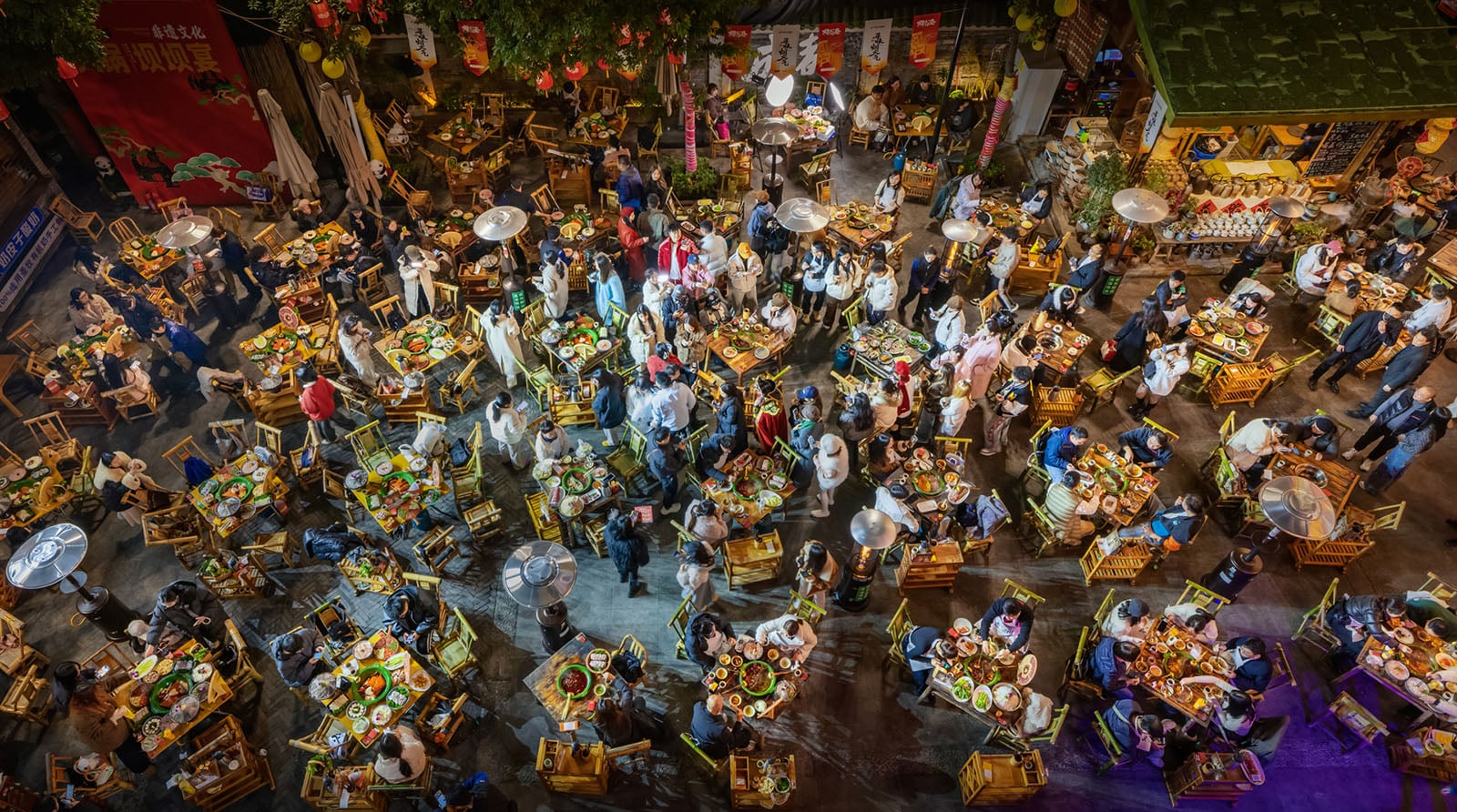 A vibrant, crowded night market with many people dining at tables filled with food, viewed from above. Warm lights and colorful dishes create a lively atmosphere in an outdoor setting.