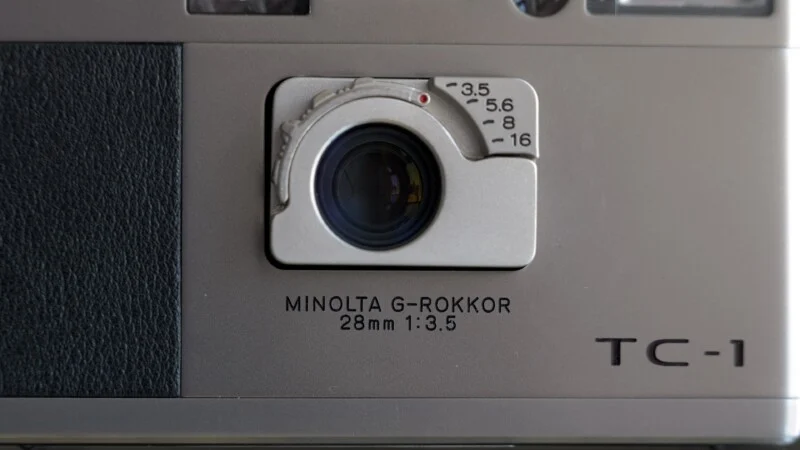 Close-up of a Minolta TC-1 camera showing the lens labeled "MINOLTA G-ROKKOR 28mm 1:3.5" with aperture settings from 3.5 to 16 visible above the lens.
