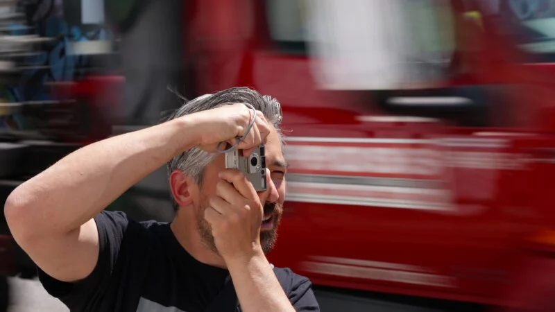 A person with gray hair is holding a camera up to their face, taking a photo. The background shows a red vehicle passing by at high speed, creating a blurred motion effect.