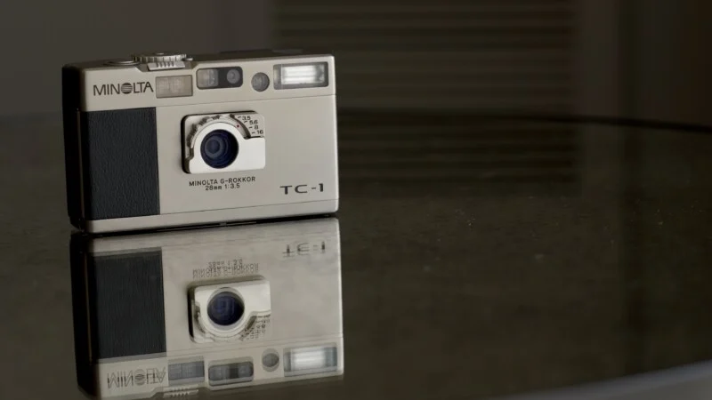 A silver Minolta TC-1 camera sits on a reflective glass surface, creating a clear mirror image beneath it. The background is softly blurred.