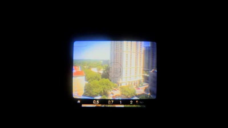 View of a cityscape with tall buildings and trees, seen through the viewfinder of a camera. The edges are dark, and camera focus markings are visible at the bottom of the frame.