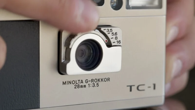A close-up of a Minolta TC-1 camera lens with a finger adjusting the aperture ring, showing f-stop numbers from 3.5 to 16 and the label "MINOLTA G-ROKKOR 28mm 1:3.5."